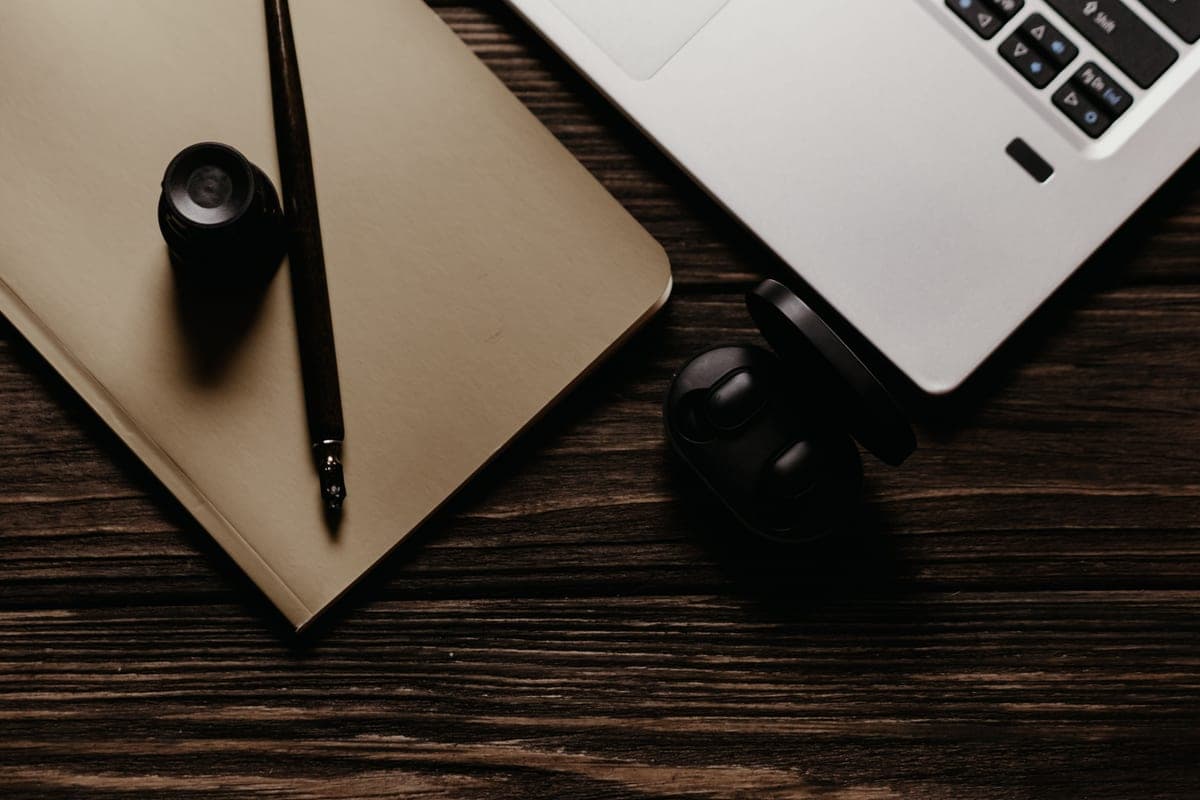 A notebook with a fountain pen and ink bottle beside a laptop on a dark wooden desk, seen from above